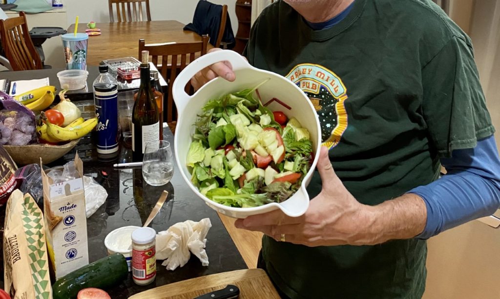 person in a home kitchen holding a homemade salad bowl with various ingredients on the counter